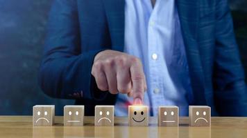 Evaluación del servicio al cliente y conceptos de calificación del servicio de la encuesta de satisfacción. La mano del hombre de negocios elige el bloque de madera con el icono de la cara sonriente de la cara feliz en el cubo de madera. foto