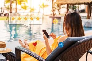 joven mujer relajante a recurso piscina y utilizando teléfono inteligente a dar un cinco estrellas satisfacción clasificación de el del hotel Servicio y habitaciones en social medios de comunicación. viaje estilo de vida concepto foto