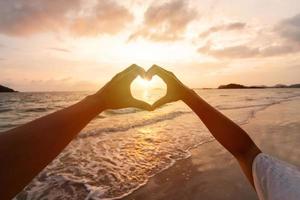 joven Pareja viajero haciendo corazón forma con manos en el playa a atardecer, amantes en Luna de miel y San Valentín día concepto foto