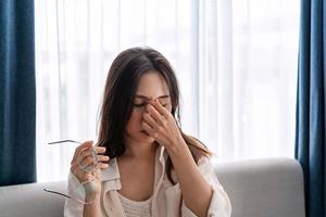 una joven que se frota los ojos siente dolor y se quita las gafas sentada en el sofá y descansa los ojos en casa, problema de visión foto