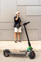 viajero diario al trabajo hipster hombre con eléctrico scooter es utilizando teléfono inteligente en el afueras foto