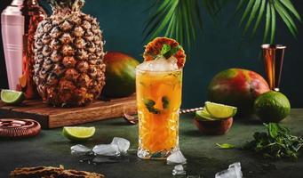Alcoholic cocktail with vodka, pineapple juice, mango, red syrup and ice. Long drink or summer cold mocktail. Tropical dark background with palm leaves and exotic fruits photo
