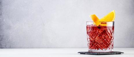 Negroni popular alcoholic cocktail with dry gin, red vermouth and red bitter, orange slice and ice cubes. Gray bar counter background, bar tools, copy space banner