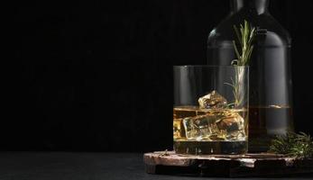 Whiskey, scotch or bourbon glass and bottle with fresh rosemary, ice cubes on black backgroun photo