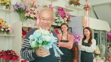 un antiguo masculino florista propietario mirando a cámara y dando hermosa ramo, hembra de los trabajadores equipo brazos cruzado juntos, contento trabajo en vistoso flor tienda almacenar, SME negocio empresario gente. video