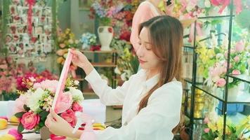 un Jeune magnifique asiatique femelle fleuriste entrepreneur organiser une bouquet de fleurs, décorer avec charmant rubans, content travail dans coloré fleur magasin boutique avec fleurit, et petit affaires propriétaire. video