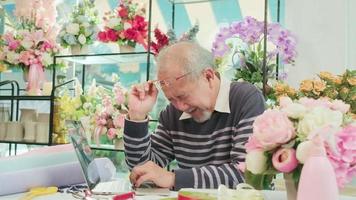 asiatisch Alten männlich Florist Inhaber ist nicht wohl mit Kopfschmerzen und Gesundheit sich verschlechtern, Stress von Überarbeitung, jung weiblich Arbeiter Unterstützung mit Pflege im ein Blume Geschäft, Familie sme Geschäft Unternehmer. video