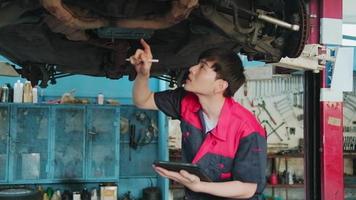 professionnel Jeune asiatique Masculin moteur mécanicien inspecte châssis de électrique voiture levé par chariot élévateur jack pour réparation à garage, automobile entretien un service travaux industrie Occupation entreprise.