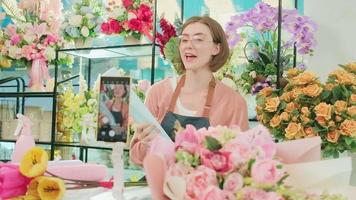 comercio electrónico negocio, uno joven blanco hembra florista demuestra y muestra floral preparativos vía en línea En Vivo transmisión con teléfono inteligente solicitud en brillante flor comercio, hermosa flores almacenar. video