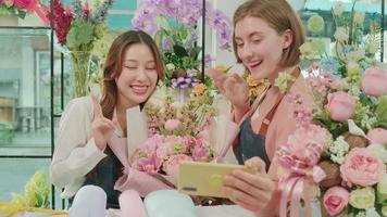 e-handel företag partner, två ung kvinna blomsterhandlare arbetare visa blommig arrangemang, uppkopplad leva strömning, och selfies med smartphone Ansökan i ljus blomma affär, skön blommar Lagra. video