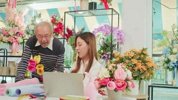 Asian young beautiful female florist worker discussing with an old man shop owner, website arrangement for online business service, happy work in colorful flower store, e-commerce SME entrepreneur.