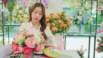 un Jeune magnifique asiatique femelle fleuriste entrepreneur organiser une bouquet de fleurs, décorer avec charmant rubans, content travail dans coloré fleur magasin boutique avec fleurit, et petit affaires propriétaire. video