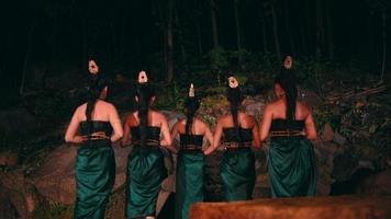 a group of Asian women standing together in front of big rocks while holding bamboo masks in their hands with flat expressions in the middle of the forest video