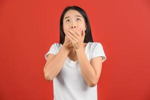 Portrait of Surprised young asian woman in white t-shirt looking up and hands cover mouth isolated on red background photo