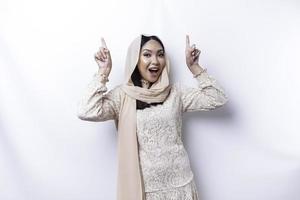 Excited Asian Muslim woman wearing hijab pointing at the copy space above her, isolated by white background photo