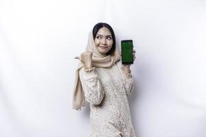 A thoughtful young Asian Muslim woman is wearing hijab and looks confused between choices while showing her phone screen, isolated by a white background photo