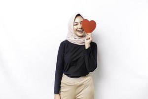 A happy young Asian Muslim woman wearing a hijab feels romantic shapes heart gesture expressing tender feelings and holding a red heart-shaped paper photo