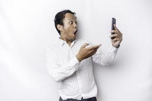 conmocionado asiático hombre vistiendo un blanco camisa y participación su teléfono, aislado por blanco antecedentes foto