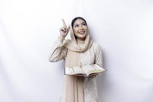 Young Asian Muslim woman smiling while pointing to copy space above her and holding the Quran photo