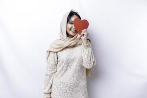 un contento joven asiático musulmán mujer vistiendo un hijab siente romántico formas corazón gesto expresando oferta sentimientos y participación un rojo en forma de corazon papel foto