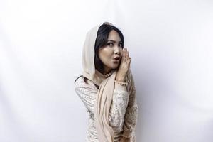 Young beautiful Asian Muslim woman wearing a hijab shouting and screaming loud with a hand on her mouth. communication concept. photo