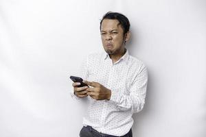 An angry young Asian man looks disgruntled wearing a white shirt irritated face expressions holding his phone photo