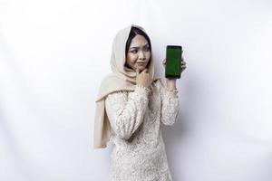 A thoughtful young Asian Muslim woman wearing a hijab and holding her chin while showing the phone screen isolated by a white background photo