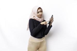 A dissatisfied young Asian Muslim woman looks disgruntled wearing hijab irritated face expressions holding her phone photo