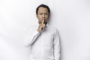 Portrait of a young Asian man tell be quiet, shushing with serious face, hush with finger pressed to lips, isolated by white background photo