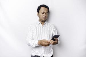 An angry young Asian man looks disgruntled wearing a white shirt irritated face expressions holding his phone photo