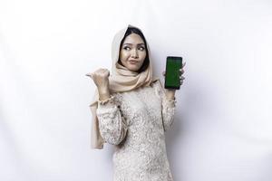 A thoughtful young Asian Muslim woman is wearing hijab and looks confused between choices while showing her phone screen, isolated by a white background photo