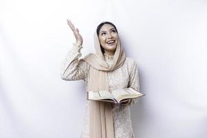 Young Asian Muslim woman smiling while pointing to copy space above her and holding the Quran photo