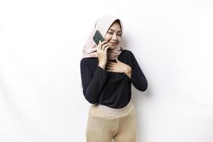 Happy mindful thankful young Asian Muslim woman with her hand on her chest smiling while talking on the phone, isolated on white background photo