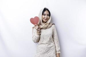 A happy young Asian Muslim woman wearing a hijab feels romantic shapes heart gesture expressing tender feelings and holding a red heart-shaped paper photo