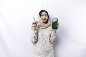 A thoughtful young Asian Muslim woman wearing hijab while showing her phone screen and pointing up to copy space above her isolated by a white background photo