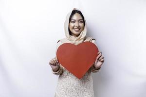 A happy young Asian Muslim woman wearing a hijab feels romantic shapes heart gesture expressing tender feelings and holding a red heart-shaped paper photo