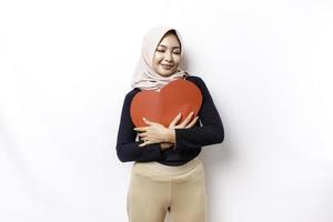 A happy young Asian Muslim woman wearing a hijab feels romantic shapes heart gesture expressing tender feelings and holding a red heart-shaped paper photo