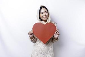 A happy young Asian Muslim woman wearing a hijab feels romantic shapes heart gesture expressing tender feelings and holding a red heart-shaped paper photo