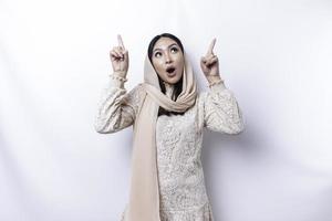 Shocked Asian Muslim woman wearing hijab pointing at the copy space on top of her, isolated by white background photo