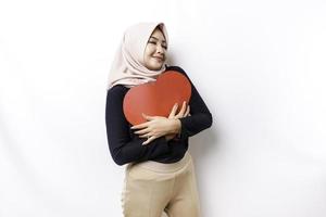 A happy young Asian Muslim woman wearing a hijab feels romantic shapes heart gesture expressing tender feelings and holding a red heart-shaped paper photo