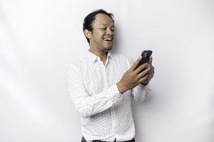 Excited Asian man wearing white shirt smiling while holding his phone, isolated by white background photo