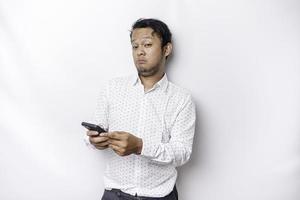 An angry young Asian man looks disgruntled wearing a white shirt irritated face expressions holding his phone photo