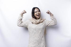 Excited Asian Muslim woman wearing a hijab showing strong gesture by lifting her arms and muscles smiling proudly photo