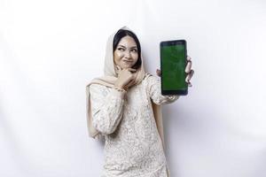 A thoughtful young Asian Muslim woman wearing a hijab and holding her chin while showing the phone screen isolated by a white background photo
