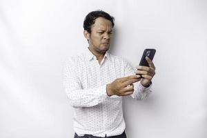 An angry young Asian man looks disgruntled wearing a white shirt irritated face expressions holding his phone photo