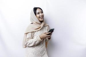A portrait of a happy Asian Muslim woman wearing a hijab, holding her phone, isolated by white background photo