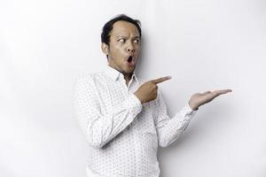 Shocked Asian man wearing white shirt pointing at the copy space beside him, isolated by white background photo