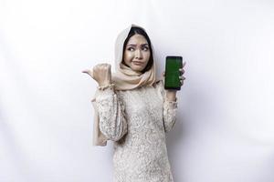 A thoughtful young Asian Muslim woman is wearing hijab and looks confused between choices while showing her phone screen, isolated by a white background photo