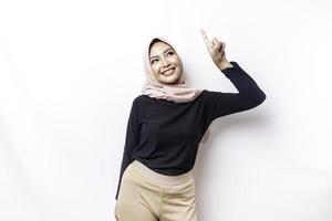 Excited Asian Muslim woman wearing hijab pointing at the copy space above her, isolated by white background photo