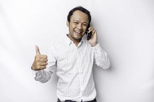 Excited Asian man wearing white shirt smiling while holding his phone, isolated by white background photo